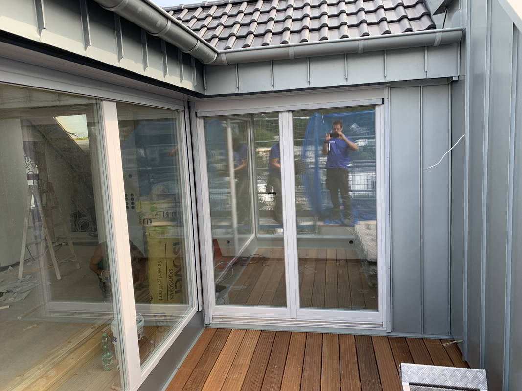 Mit Bankirai Holz belegte Dachterrasse. Man sieht zwei große Glasschiebetüren und die Seitenverkleidung der Fassade aus Zinkblech.